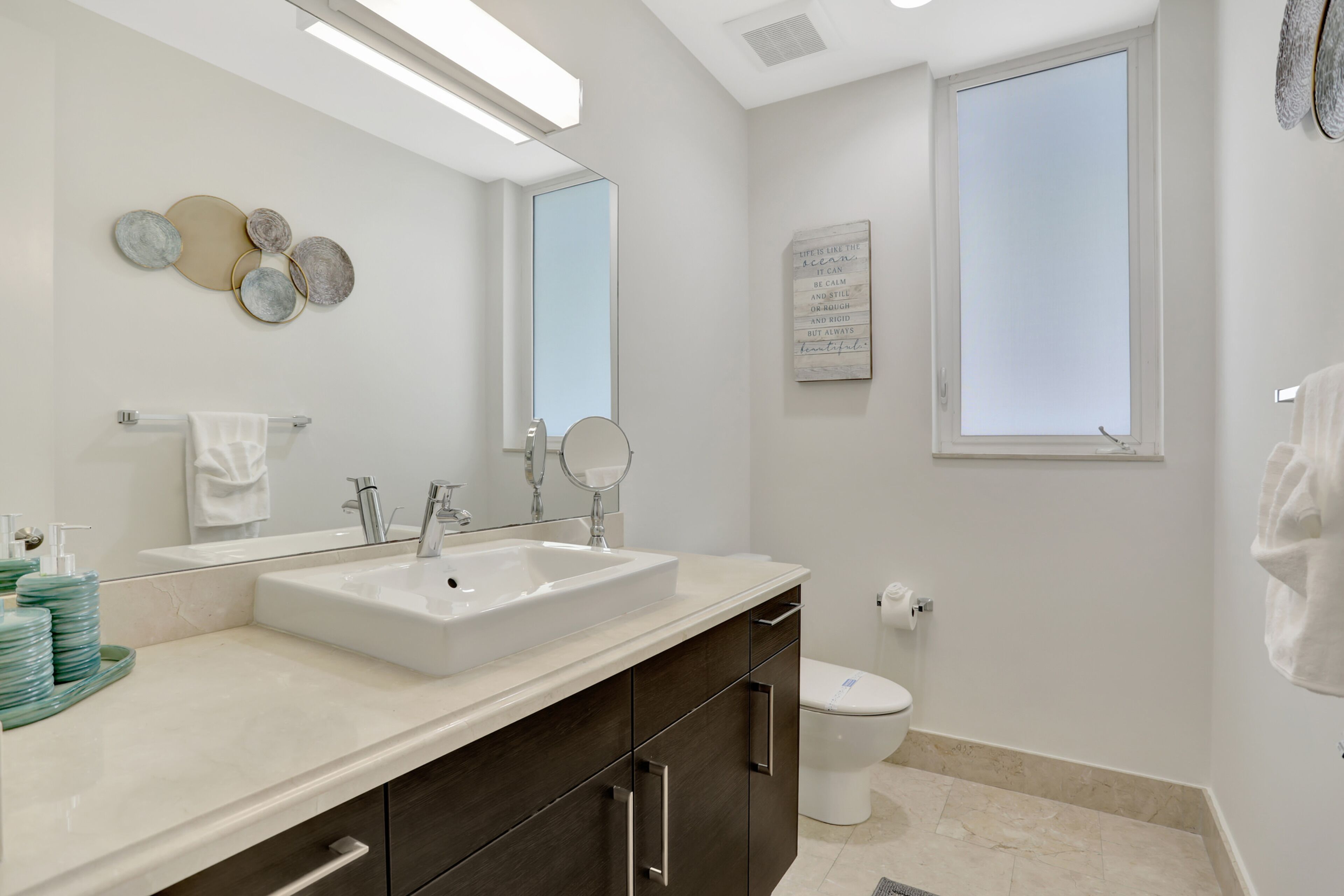 A modern bathroom with a large sink, dark cabinets, a toilet, and a window providing natural light.