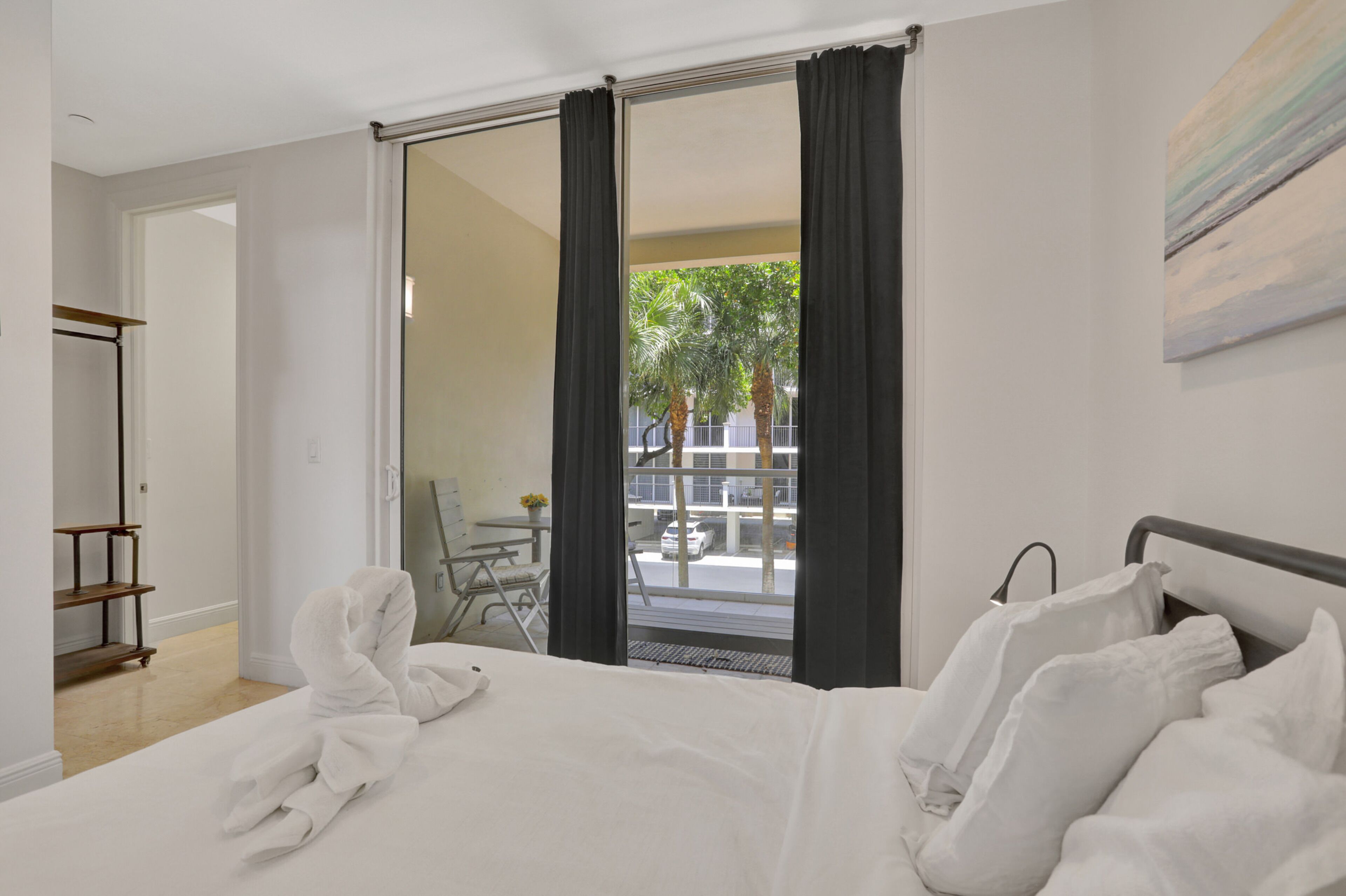 Une chambre d’hôtel avec un lit, un bureau et une chaise. Elle dispose d’un balcon avec vue sur des palmiers et un immeuble.