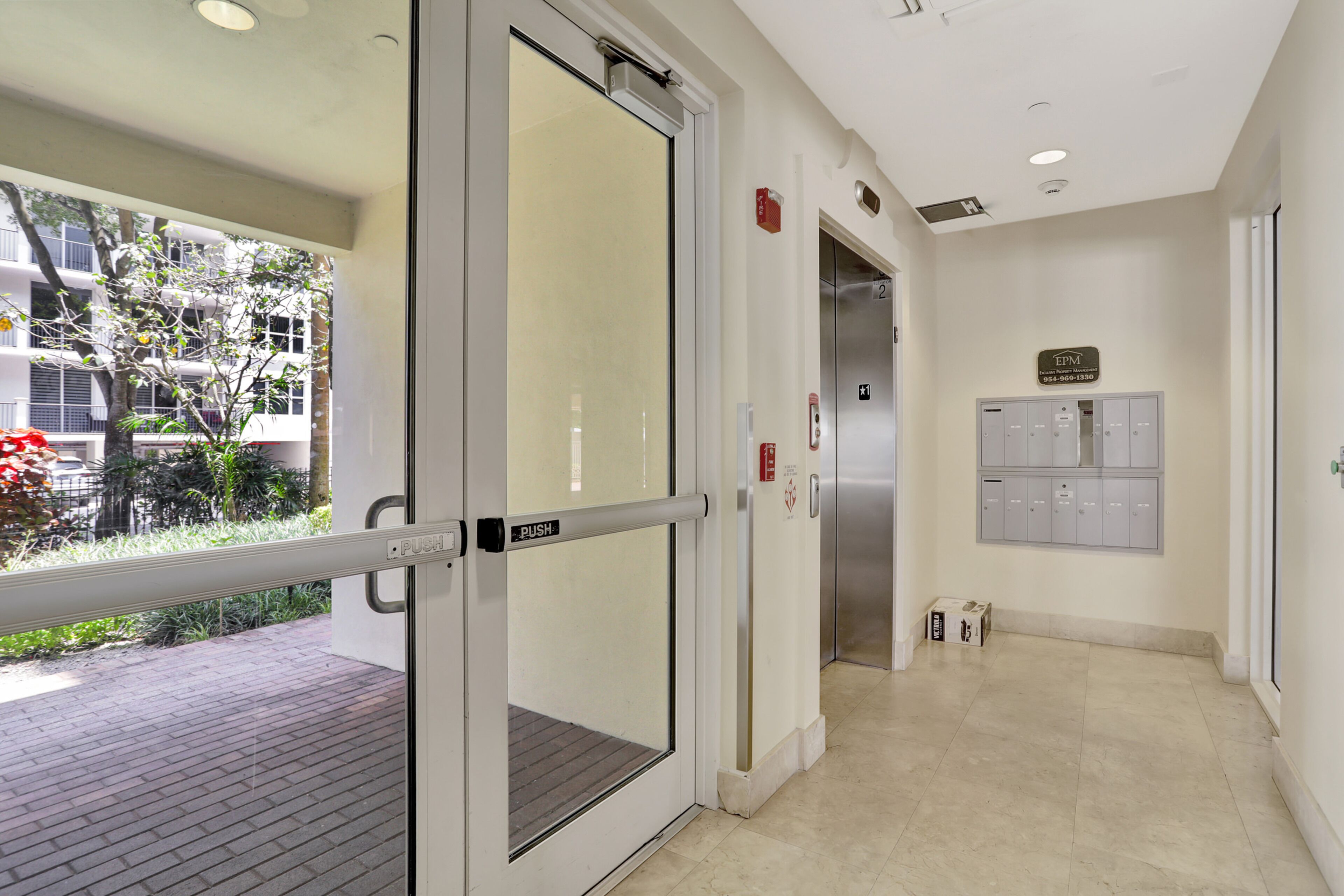 L’entrée d’un hôtel, avec une porte en verre, une barre de poussée, un détecteur d’incendie, un ascenseur et une rangée de casiers.