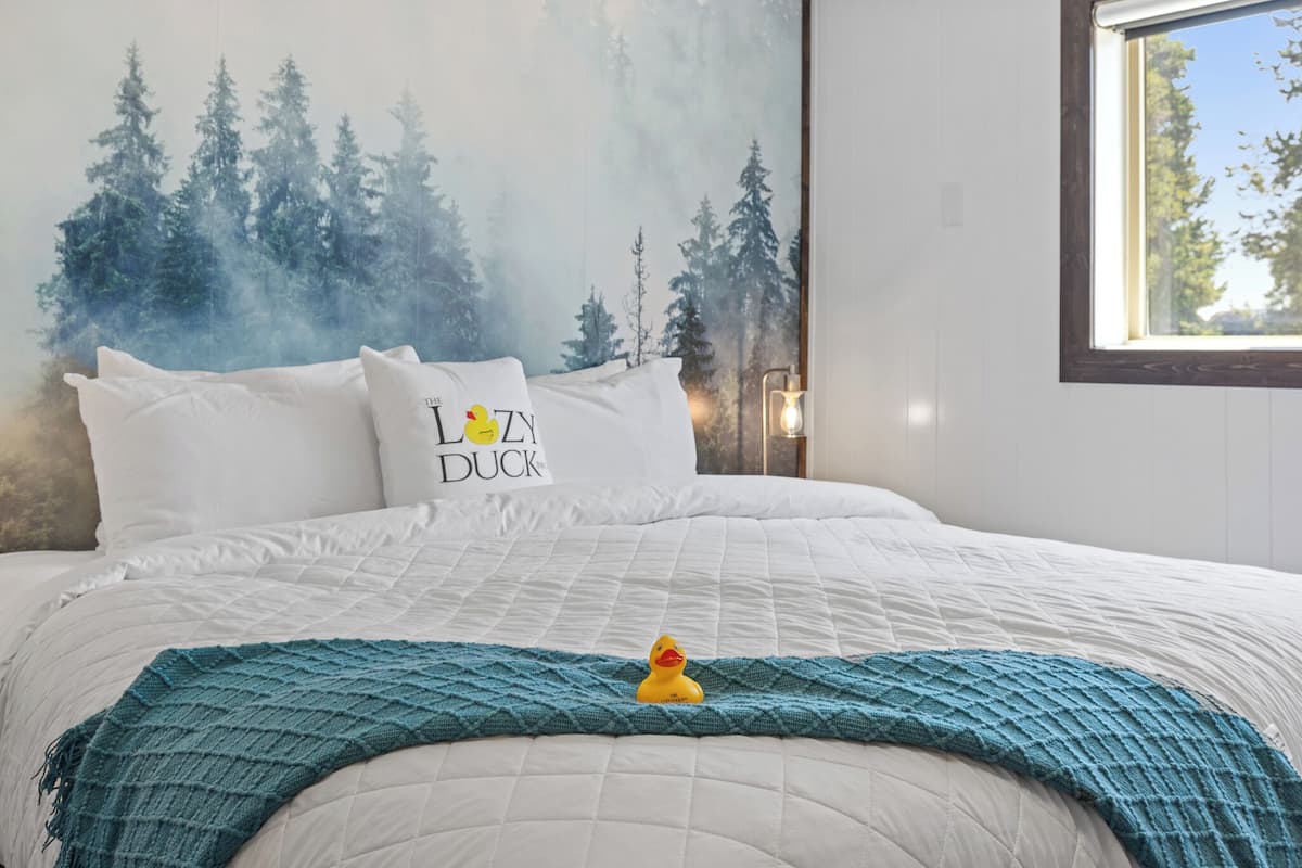 Un lit bien fait, recouvert d’une couette blanche, d’un plaid turquoise et d’un oreiller orné d’un motif de canard.