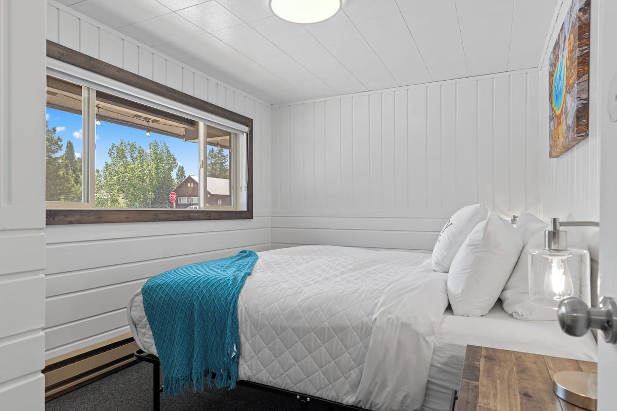 Une chambre à coucher avec un lit, une fenêtre donnant sur des arbres et un immeuble, et un tableau accroché au mur.