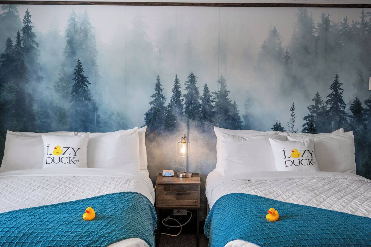 Une chambre d’hôtel avec deux lits, chacun agrémenté d’un oreiller orné du logo de « The Lazy Duck Inn », une table de chevet avec une lampe et une fresque murale sur le thème de la forêt.