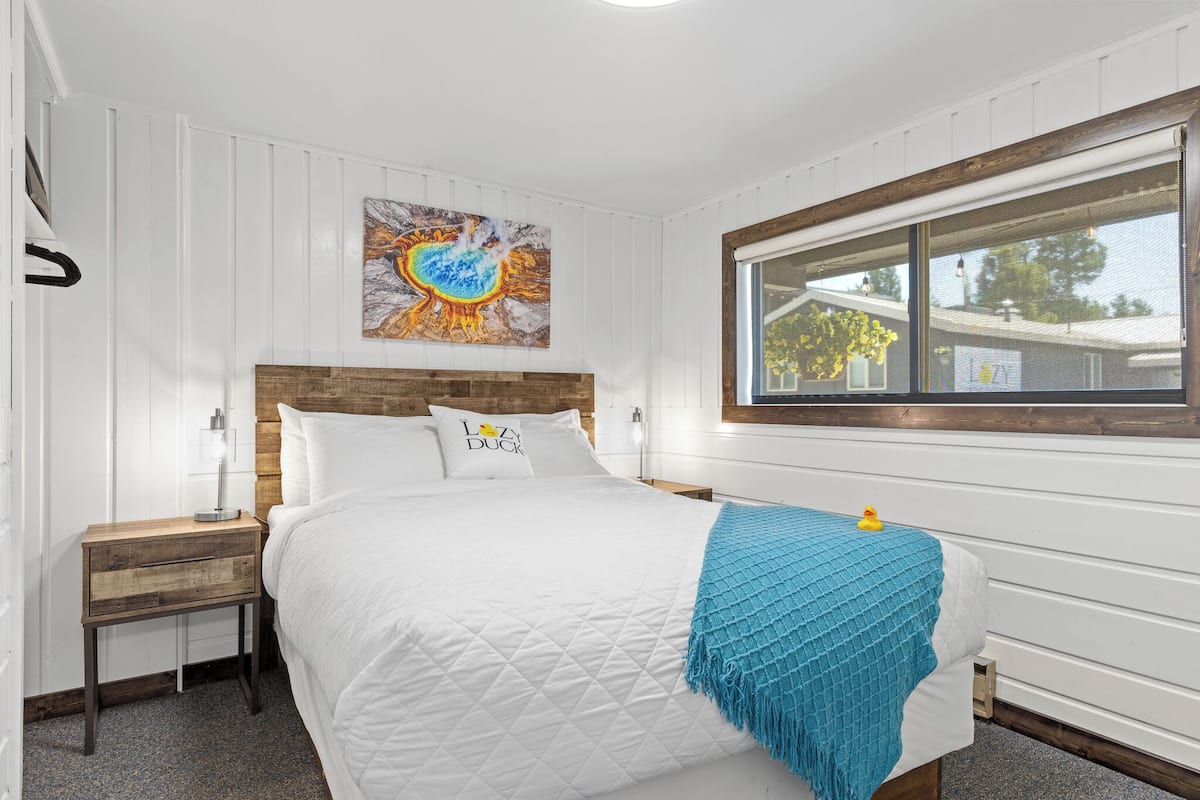 Une chambre à coucher avec une tête de lit en bois, du linge de lit blanc, un plaid bleu, une table de chevet avec une lampe et une fenêtre donnant sur l’extérieur.