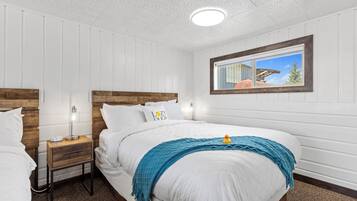 A bedroom with two beds, wooden headboards, white walls, a window with a view, and a nightstand with a lamp.