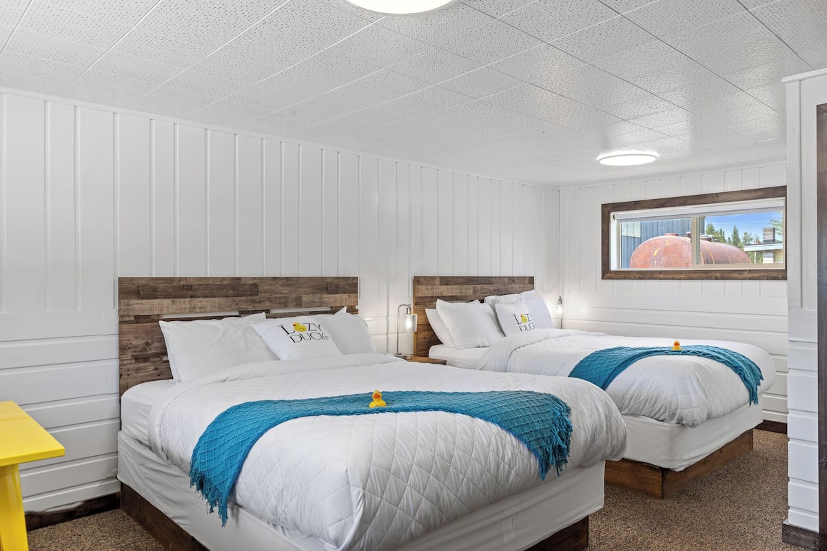Une chambre d’hôtel avec deux lits, du linge de lit blanc, une tête de lit en bois, une table de chevet jaune et une fenêtre donnant sur un immeuble.