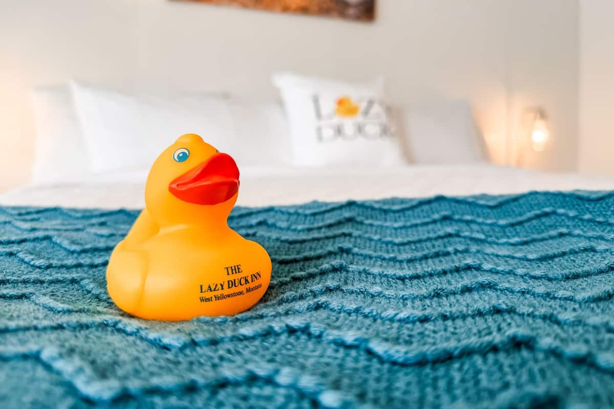Un lit avec une couverture bleue à motifs, un oreiller blanc orné de texte et un canard en caoutchouc jaune posé sur la couverture.