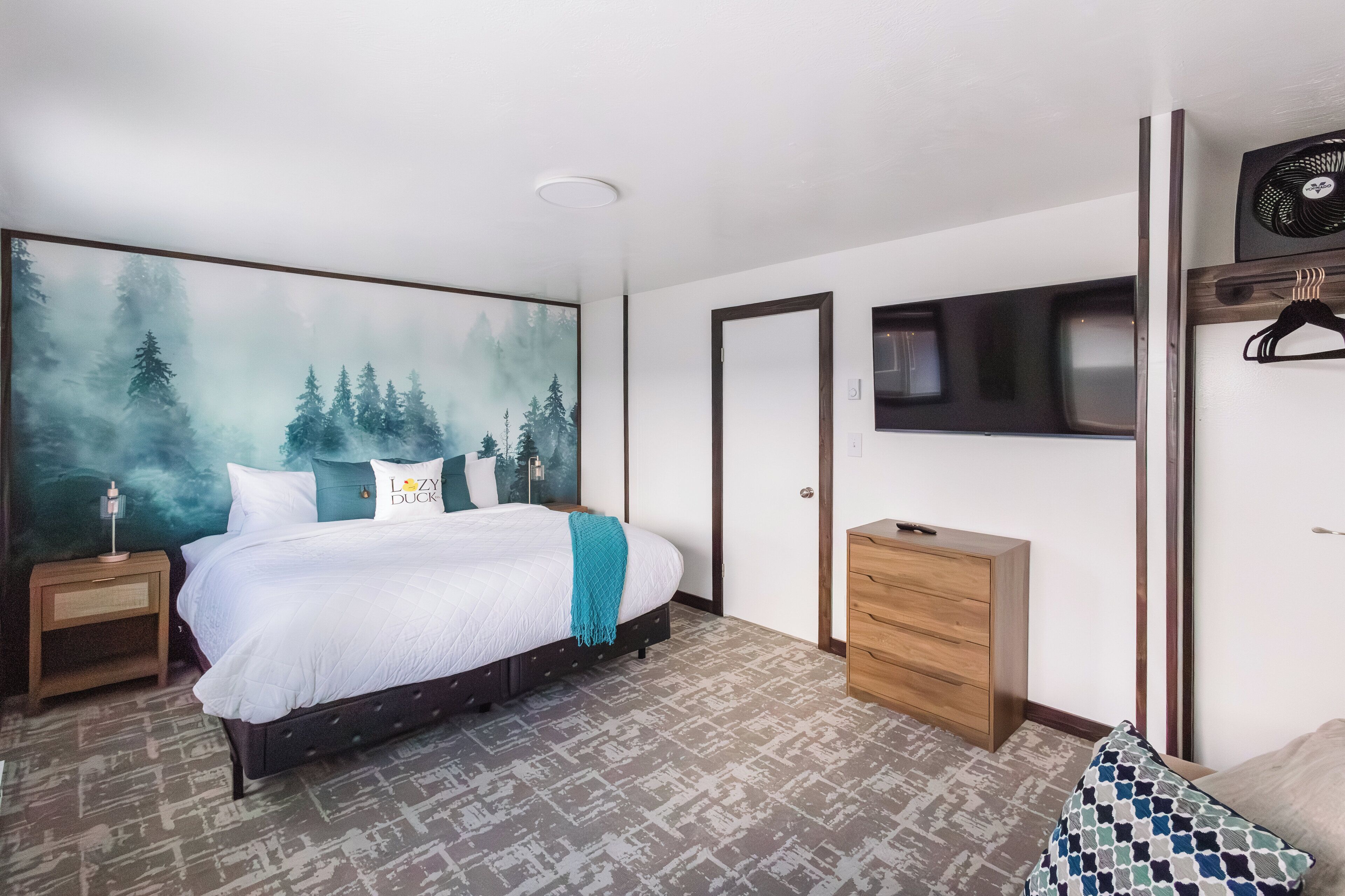 Une chambre à coucher comprenant un lit, une commode, un téléviseur fixé au mur et un grand tableau représentant une forêt.