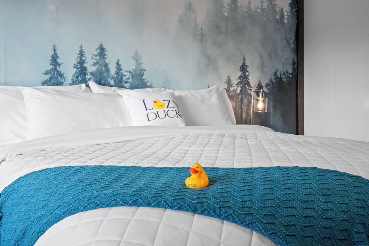 Un lit avec un matelas matelassé blanc, un plaid bleu tricoté et un canard en caoutchouc jaune. Un oreiller avec l’inscription « The Loo Duck » est visible.