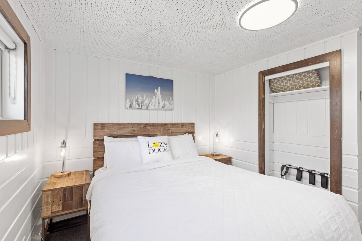 Une chambre à coucher bien rangée, avec une tête de lit en bois, du linge de lit blanc et un tableau accroché au mur.