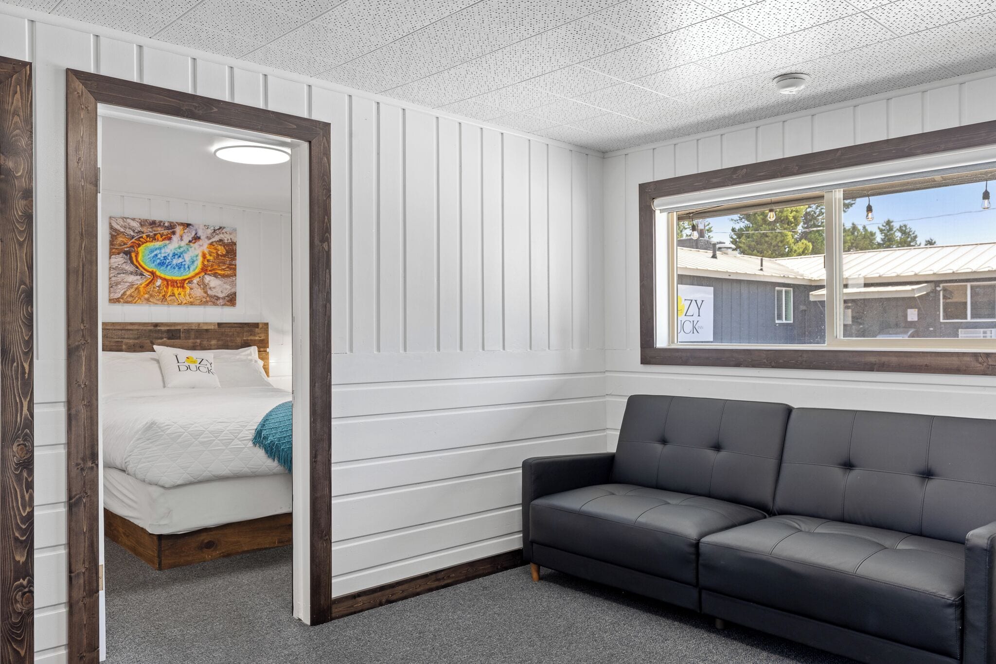 A room with a bed, a grey sofa, a window with a view of a building, and a ceiling with a grid pattern.