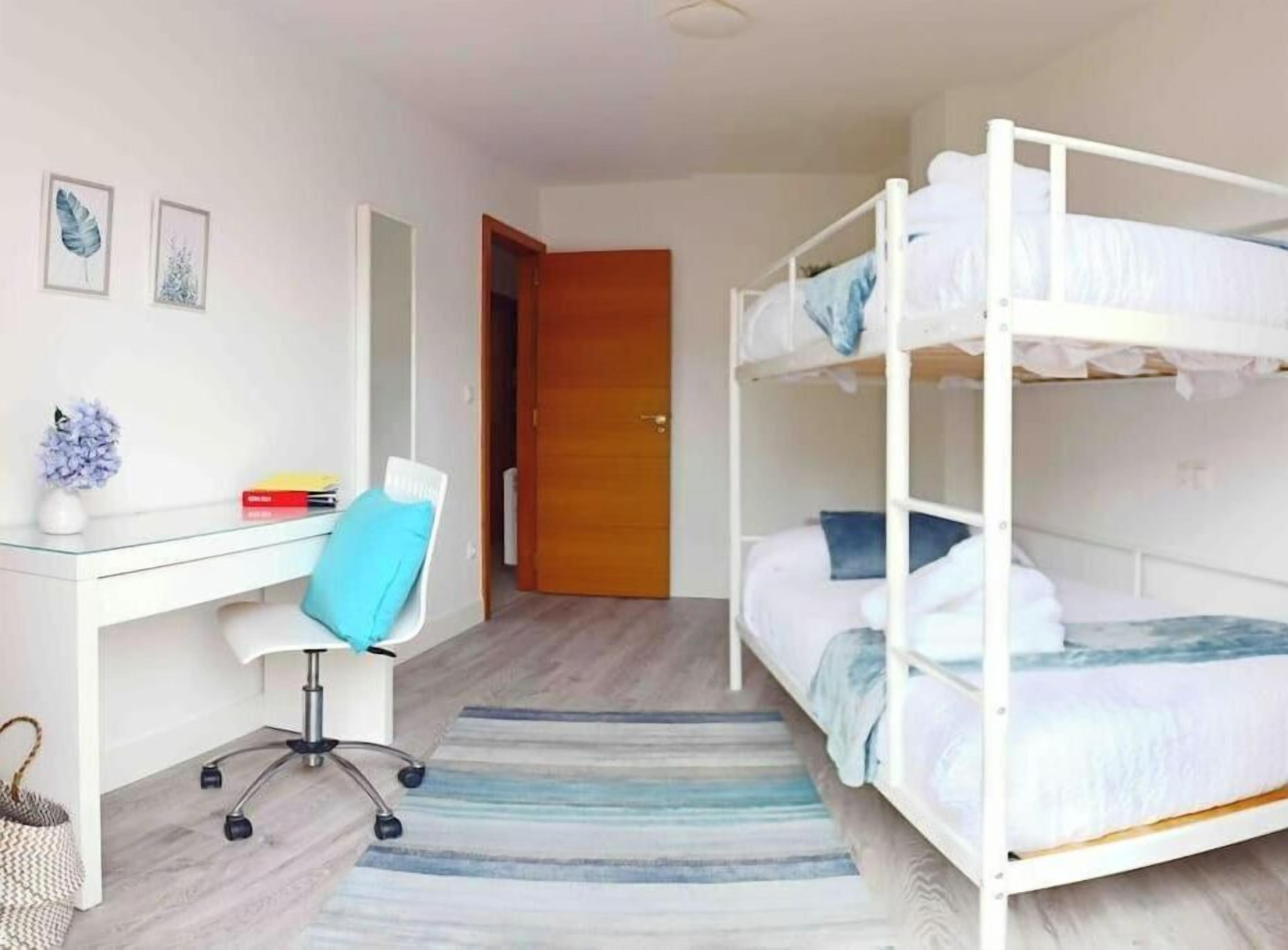 A room with a bunk bed, a desk with a chair, a small table with a vase, and a door leading to another room.