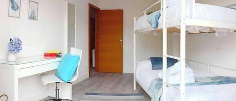 A room with a bunk bed, a desk with a chair, a small table with a vase, and a door leading to another room.