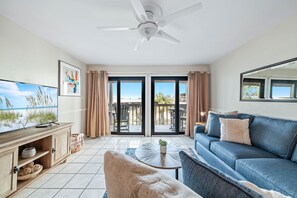 A living room with a blue sofa, a television, a ceiling fan, and a view of the outdoors through large sliding doors.