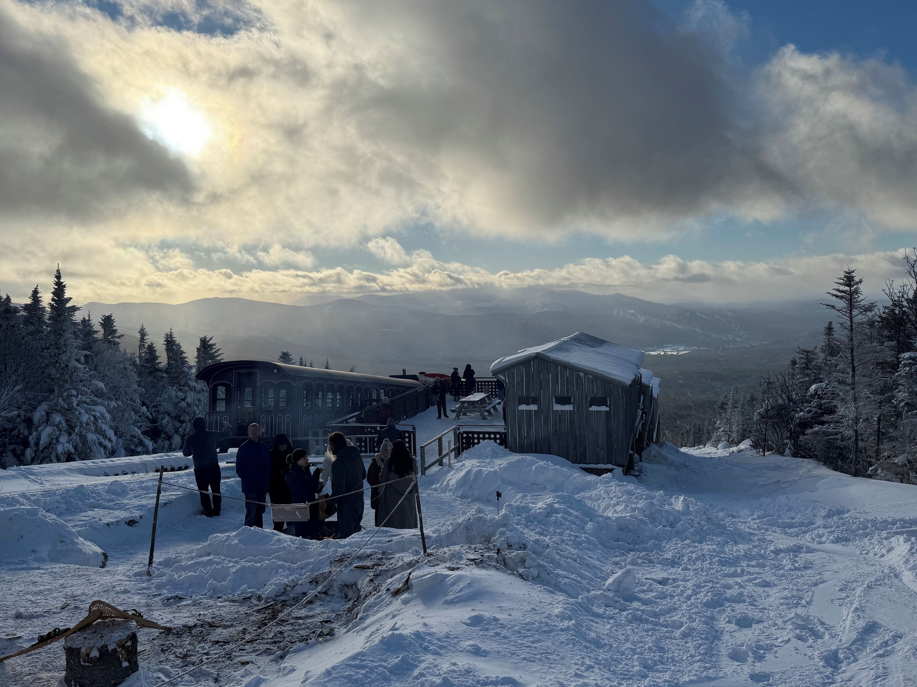A wooden cabin with a snow-covered roof, surrounded by snow-covered trees and a mountainous backdrop.