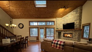 A cozy living room with a stone fireplace, wooden ceiling, and large windows overlooking a deck.