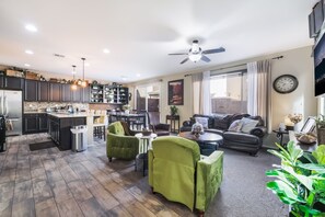 A modern kitchen with dark cabinets, a dining area with chairs, a living room with a sofa and green armchairs, and a ceiling fan.