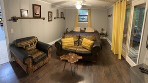 A cozy living room with a sofa, armchair, and coffee table. There is a window with curtains, a ceiling fan, and a door leading to another room. - New Guest suite located in Jasper with a mountian view and fire pit (Jasper)