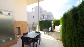A patio with a table and chairs, a grill, and a hedge.