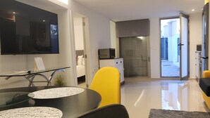 A modern living room with a flat-screen TV mounted on the wall, a dining area with a glass table and chairs, and a kitchen area with a microwave and cabinets.