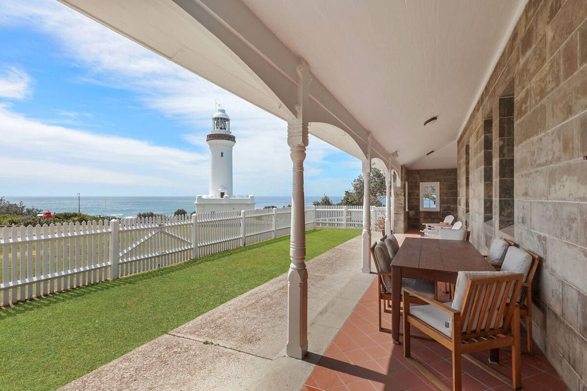 A covered patio with a view of a lighthouse, a white fence, and a grassy area.