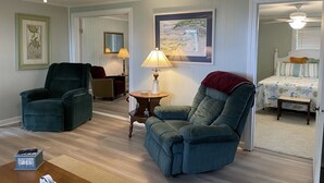 A living room with two armchairs, a coffee table, a lamp, a ceiling fan, a framed picture, and a door leading to a bedroom.