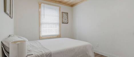 A bedroom with a bed, a window with blinds, a bedside table with a lamp, and a framed picture on the wall.