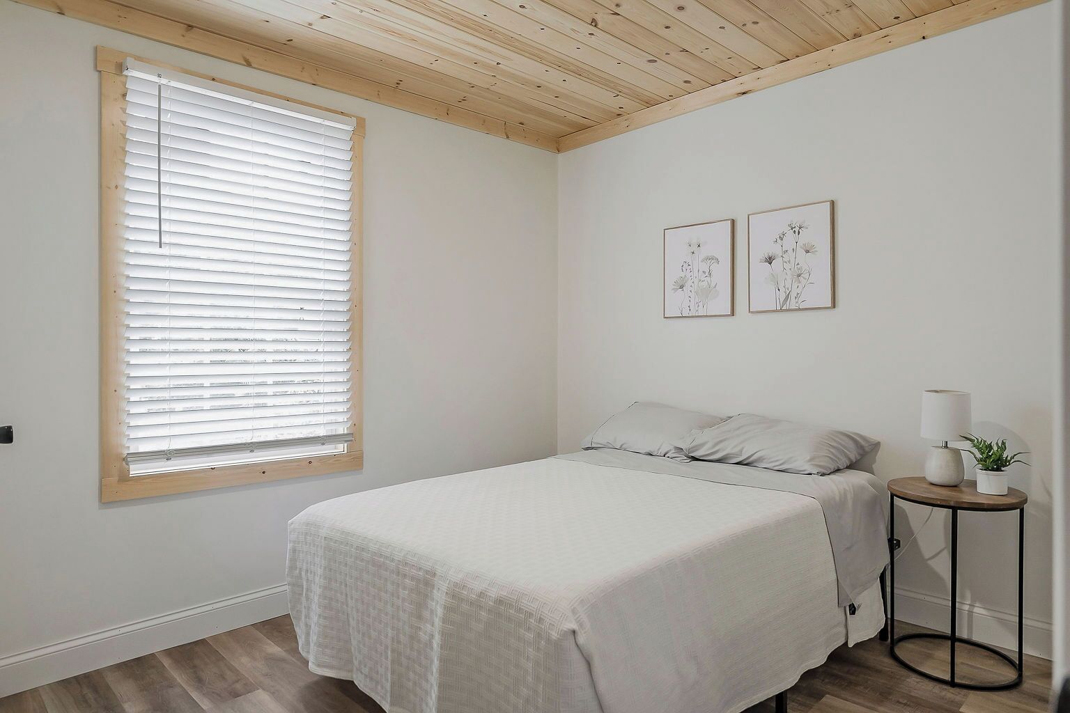 Un dormitorio con cama, ventana con persianas, mesita de noche con lámpara y dos cuadros en la pared.