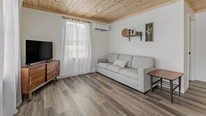 Una sala de estar con techo de madera, un televisor de pantalla plana sobre un mueble de madera, un sofá blanco, una mesita y una ventana con cortinas traslúcidas.