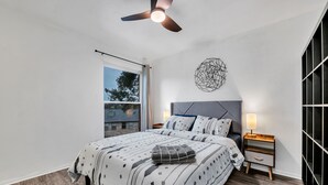 Une chambre à coucher comprenant un lit, une table de chevet, une lampe fixée au mur, un ventilateur de plafond et une fenêtre donnant sur des immeubles.