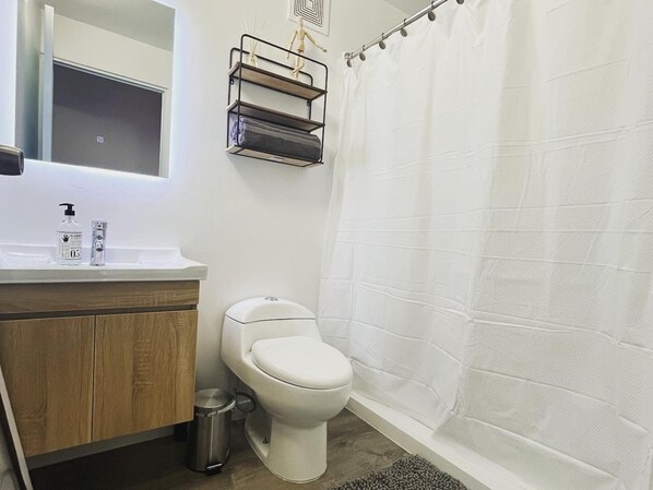 Et badeværelse med toilet, håndvask med spejl, en hylde med en pynteting og en bruseforhæng.