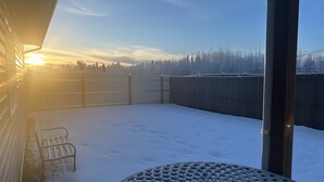 A snow-covered patio with a metal table and a bench, overlooking a wooden fence and a forested area. - Experience Fairbanks comfortably, just a 3-min drive from Creamer's Field (Fairbanks)