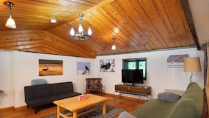 A cozy living room with a wooden ceiling, a black sofa, a green couch, a wooden coffee table, and a television on a stand.