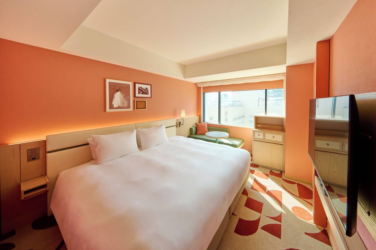A hotel room with a large bed, a television, a desk with a chair, and a window with a view of buildings.