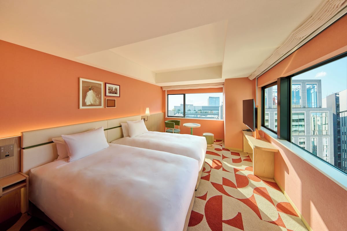 A hotel room with two beds, a small table, a chair, a window with a city view, and a wall-mounted TV.