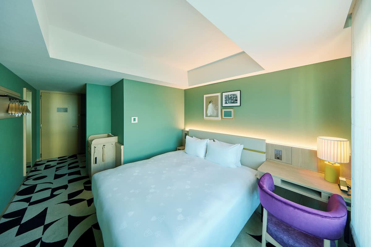 A hotel room with a bed, a desk, a chair, a lamp, and a door.
