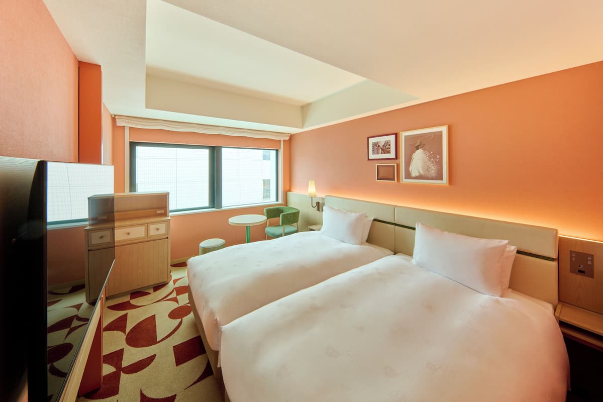 A hotel room with two beds, a desk, a chair, a small table, and a window.