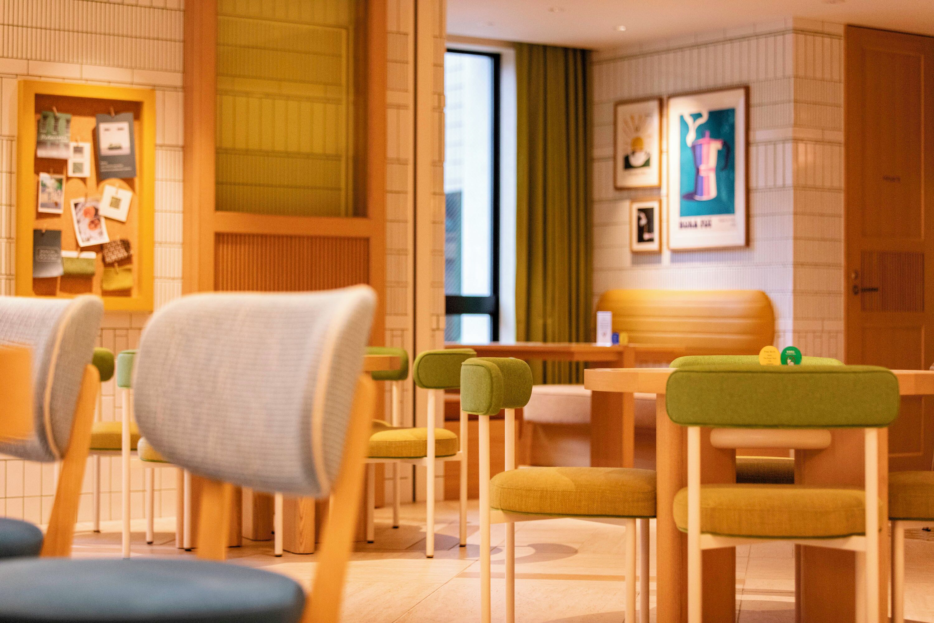 A modern hotel room with a wooden reception desk, green and yellow chairs, a large window with curtains, and framed artwork on the wall.