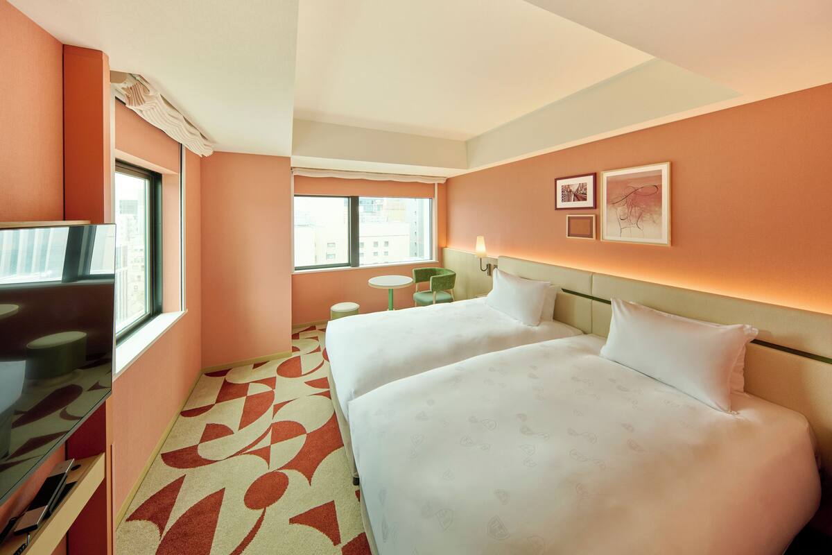A hotel room with two beds, a small table, a chair, and a window.