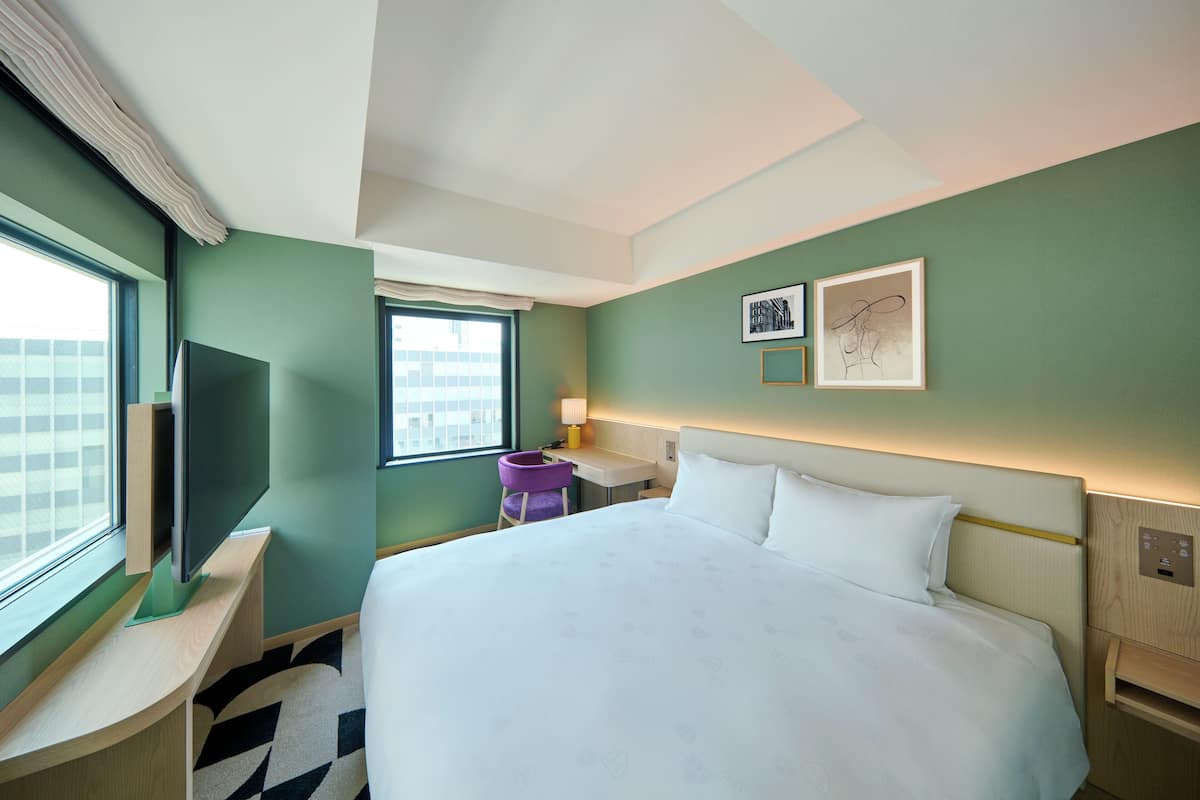 A hotel room with a large bed, a desk with a flat-screen TV, a small purple chair, and two framed pictures on the wall.