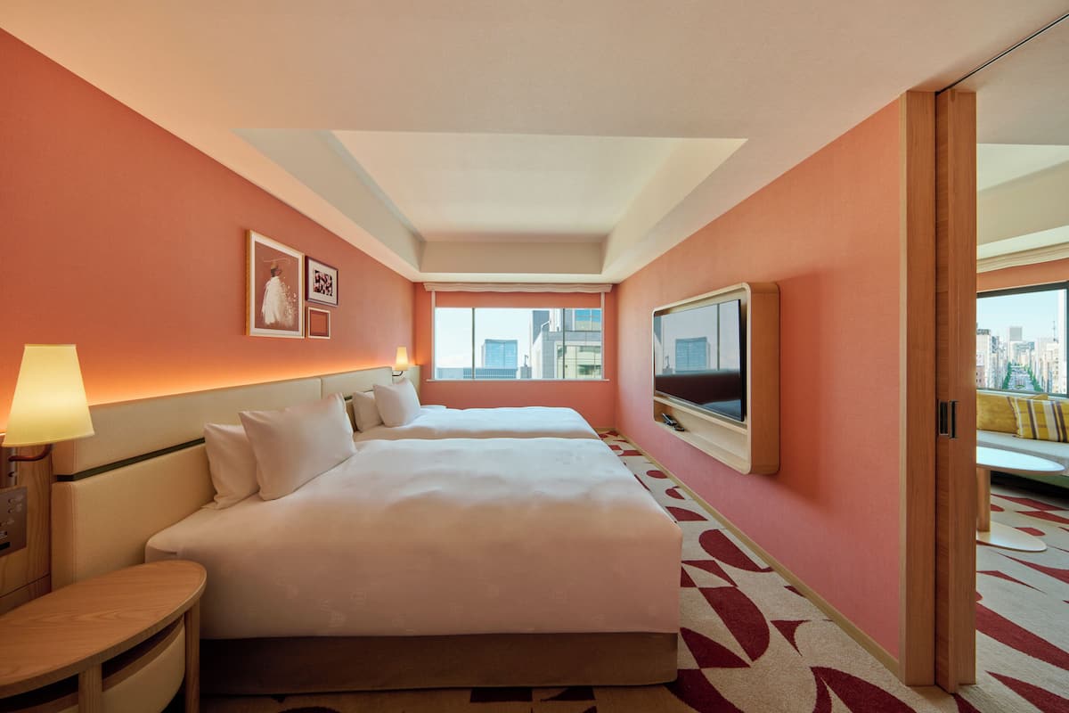 A hotel room with two beds, a TV, a window with a city view, and a patterned carpet.
