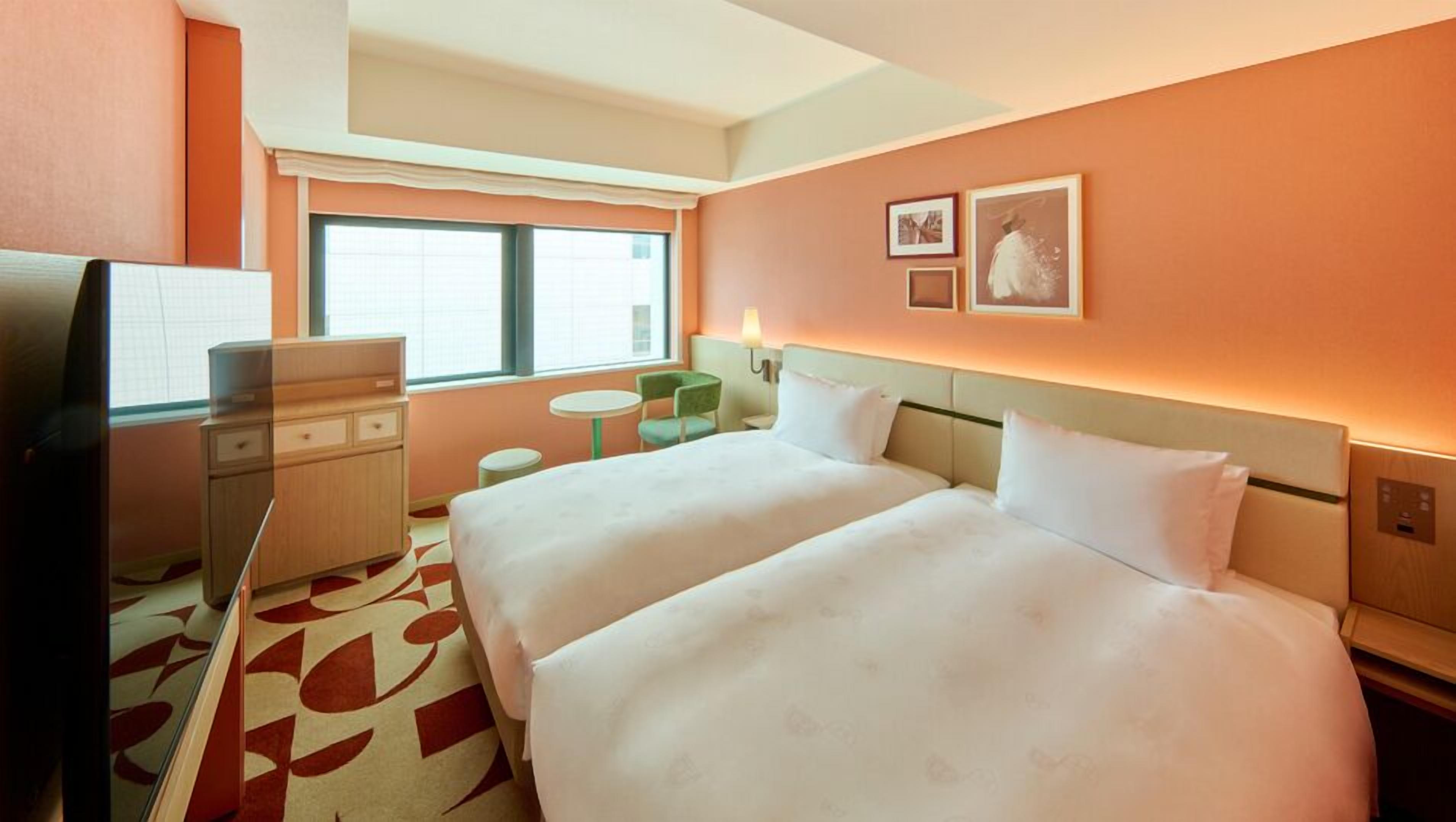 A hotel room with two beds, a desk, a chair, a TV, and a window with blinds.