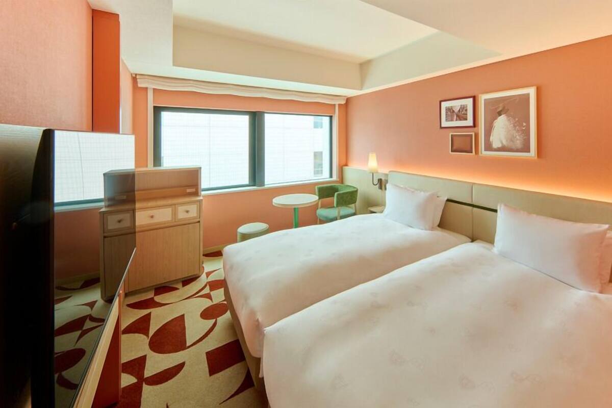 A hotel room with two beds, a desk, a chair, a TV, and a window with blinds.