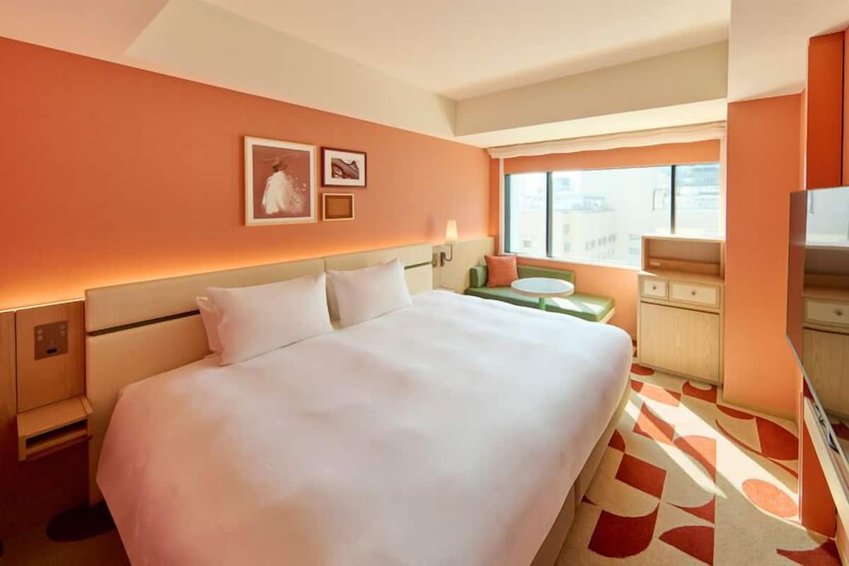 A hotel room with a large bed, a television, a small table, and a window with a city view.