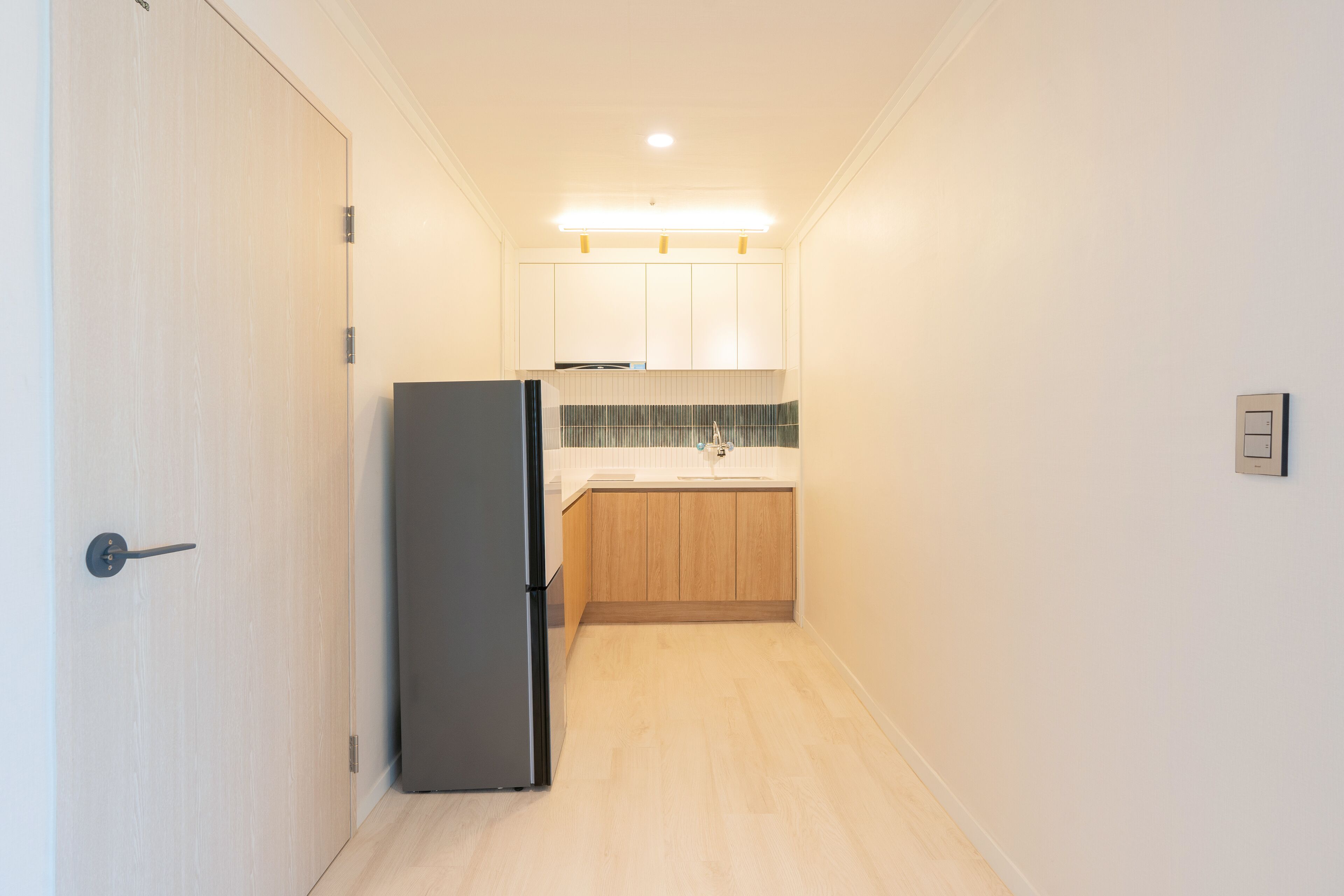A narrow hallway leading to a kitchen with wooden cabinets, a refrigerator, and a tiled backsplash.