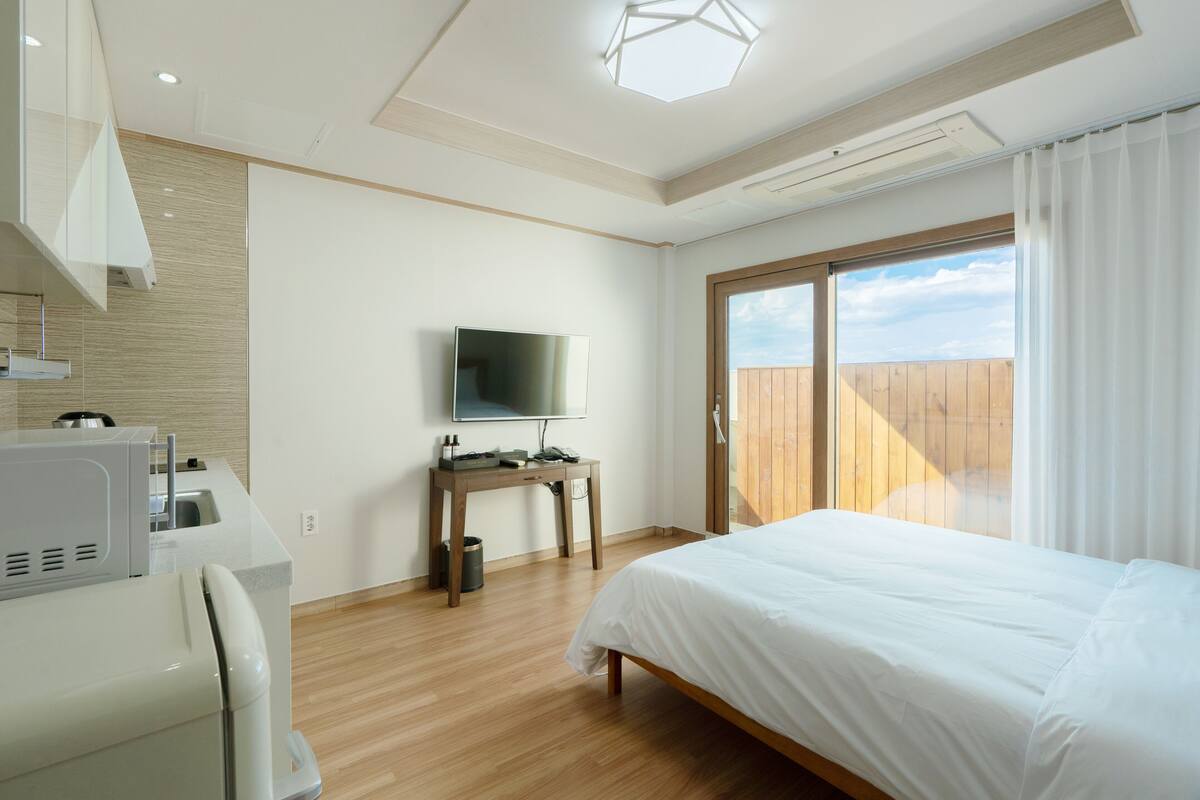 Moderni hotellihuone, jossa on puulattia, valkoisilla tekstiileillä varustettu sänky, litteänäyttöinen televisio puupöydällä ja suuri ikkuna, josta avautuu näkymä taivaalle.