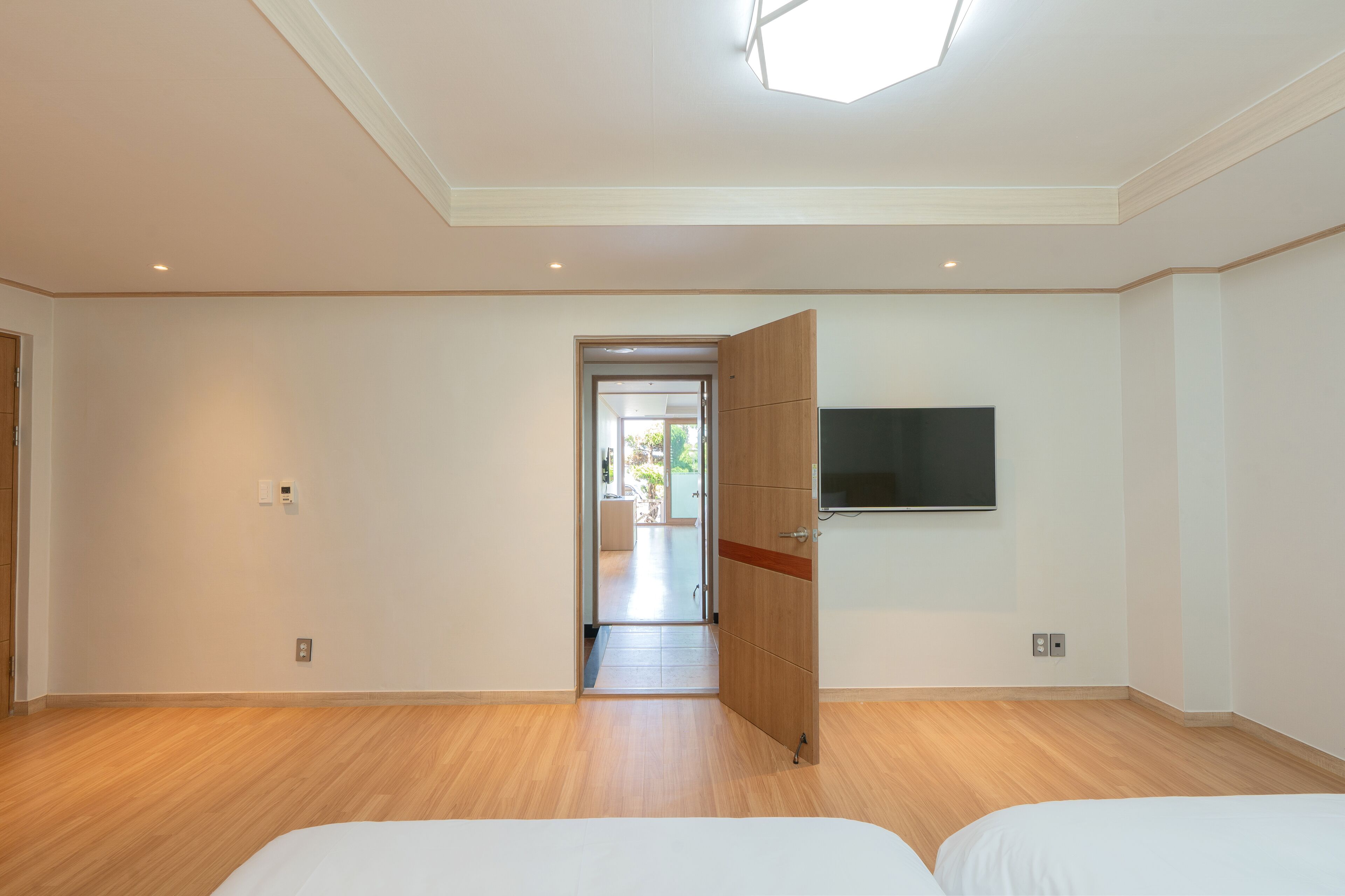 A modern hotel room with a wooden floor, a bed, a TV mounted on the wall, and a door leading to a balcony.