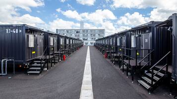 A row of identical, dark-colored, modular hotel rooms with numbered doors, a central walkway, and a building with a tall, rectangular structure in the background.
