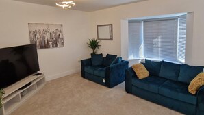 A modern living room with a flat-screen TV, two dark blue sofas, a large window with blinds, and a cityscape painting on the wall. - Peterborough - House - Sleeps 8 - Parking - Garden (Peterborough)