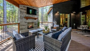 Une terrasse en bois avec des meubles en osier, une cheminée en pierre et un téléviseur fixé au mur.