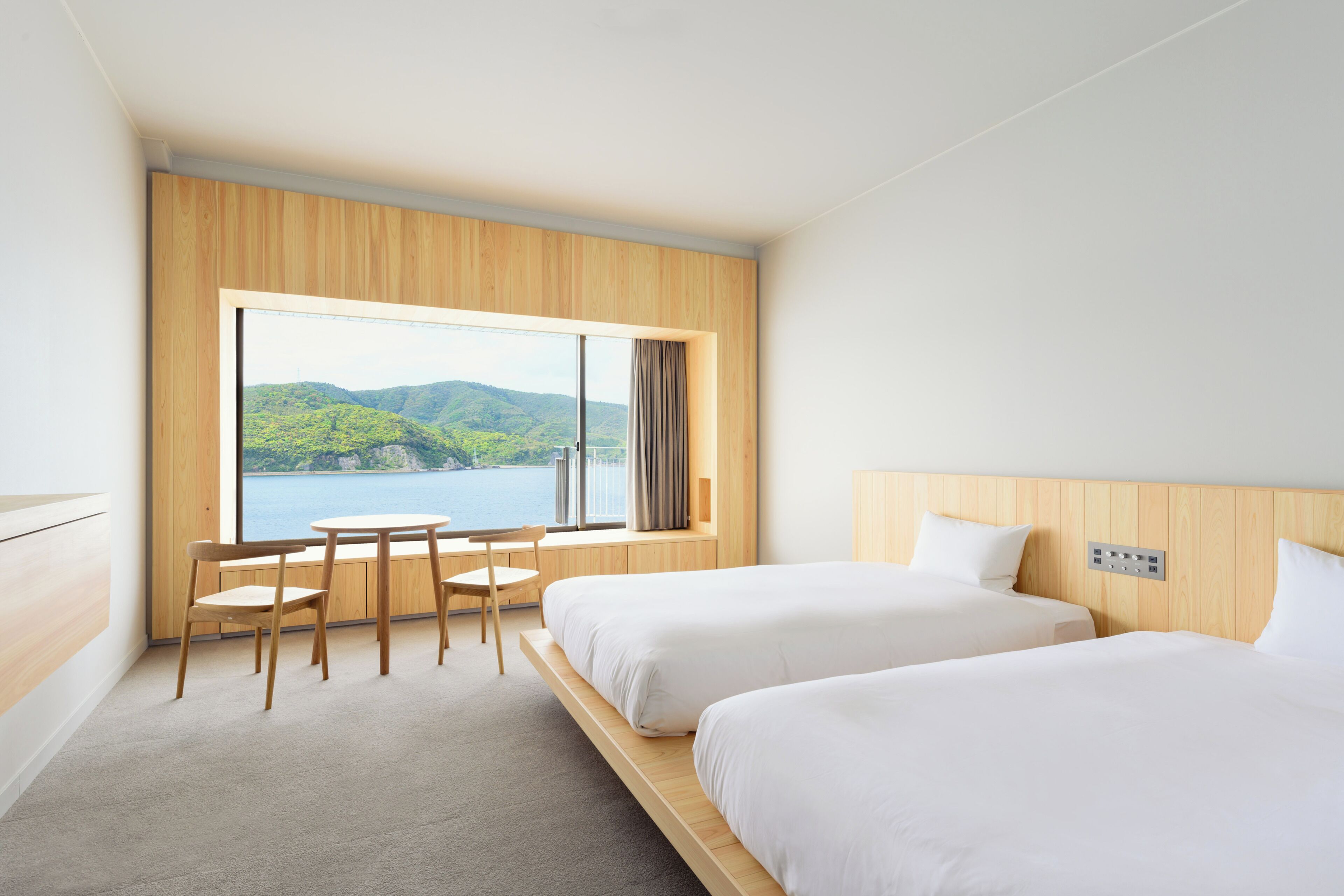 Habitación de hotel con dos camas, cabecero de madera, un ventanal con vistas a un lago y montañas, una pequeña mesa con una silla, y piso alfombrado.